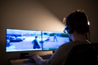 A teenage boy sits in the dark, headset on, in front of a dual computer screen that has Fortnite on the screen