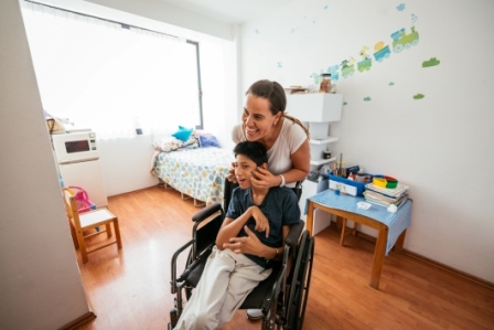 A teen boy with cerebral palsy in a wheelchair with his mom hugging him from behind