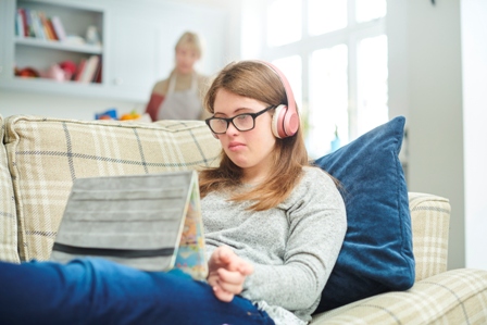 A teenage girl with down's syndrome sits on the couch looking at her laptop screen while a caregiver is in the background
