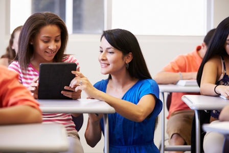 A teacher helps a teenage black girl at her desk in a classroom