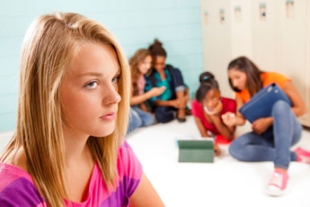 A teenage girl looks upset as a group of girls sit behind her, laughing, while looking at device screens