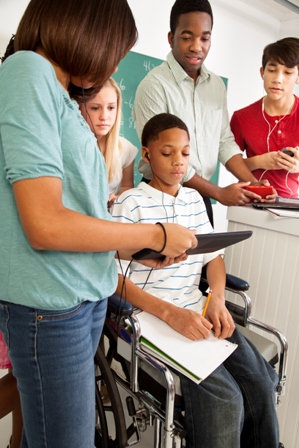 A group of students, including a boy in a wheelchair, listen to their teacher who is holding an ipad