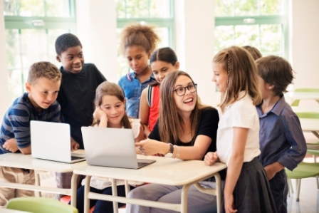 A group of diverse young students stand around their teacher who is working on a laptop