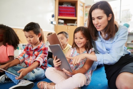 A woman teacher with a diverse group of young children looks at an ipad