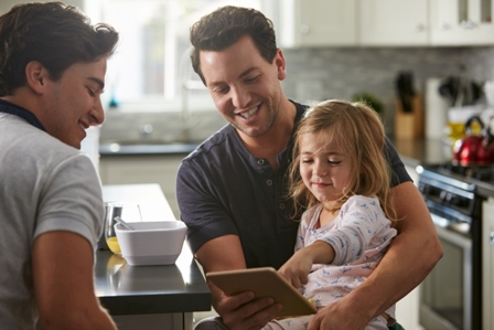 A same sex male couple, with their young daughter, sit at the kitchen island looking at a tablet screen