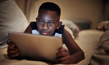 An African American boy lays on his bed, looking at an iPad screen
