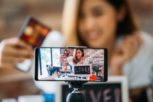 A photo of a smartphone screen, with the focus on the screen as it films a teen girl in the background doing a makeup tutorial